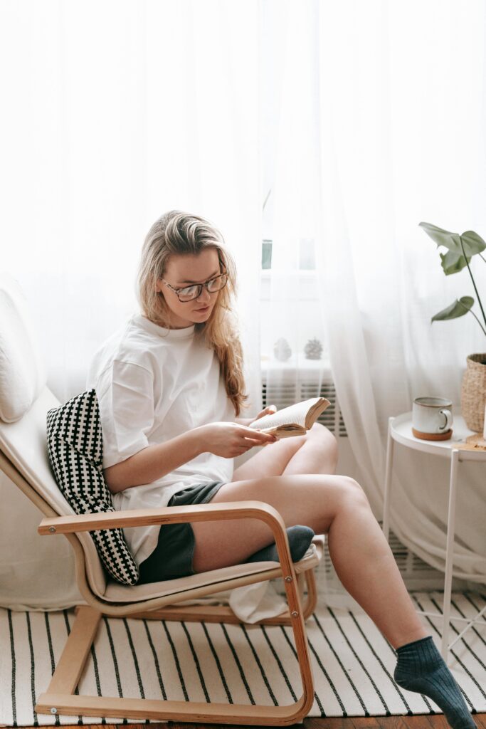 Full body of serious female in home wear sitting on armchair with cushion and reading book at home